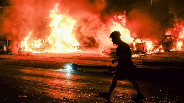 美国威斯康辛州基诺沙市一名男子走过被示威者纵火焚烧的汽车（25/8/2020）