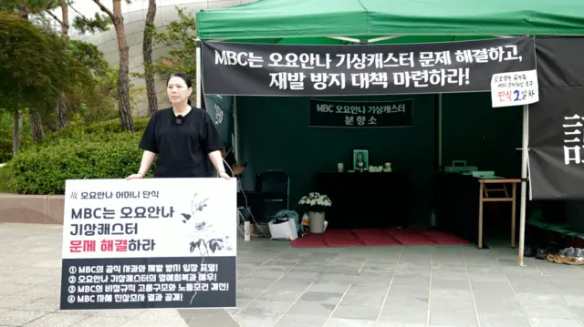 오요안나 기상캐스터의 어머니 장연미 씨가 'MBC는 오요안나 기상캐스터 문제 해결하라'는 팻말을 들고 MBC 앞에서 단식 농성을 하고 있다. 그의 뒤에는 분향소가 차려져 있다. 