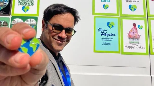 Saman Jamshidifard, embajador de Hearty Earth, sonríe frente a una pared blanca con carteles que muestran diferentes ofertas de Hearty Earth, como Happy Cakes. Muestra un pin con forma de corazón que representa la Tierra.