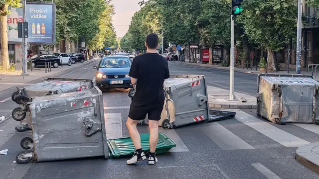 Momak okrenut leđima stoji na ulici ispred kontejnera i gleda u automobile