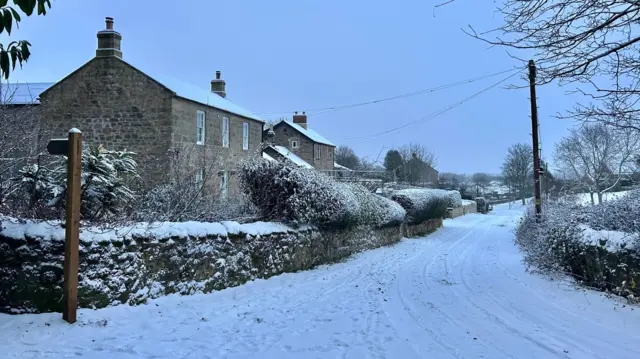 Casas de piedra a la izquierda con techos nevados. Frente a ellas hay un muro de piedra con setos y plantas que las cubren. El camino frente a las propiedades tiene algunas huellas de neumáticos, mientras que el cielo se tiñe de azul. Presenta una pintoresca escena de un pueblo nevado.