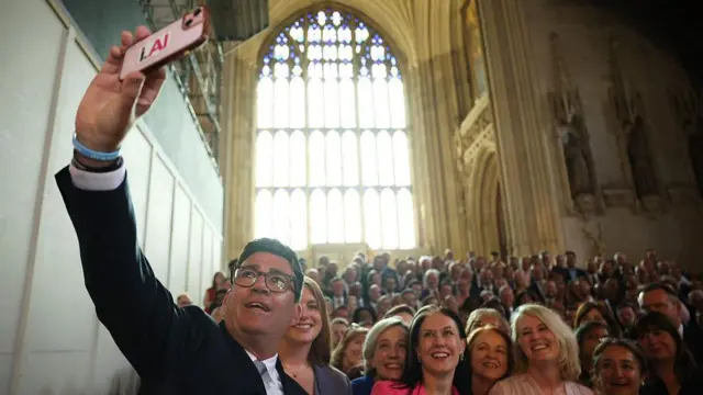 英議会議事堂のゴシック様式の壁や彫像や窓を背に、階段に並ぶ数十人の前で、スマートフォンで自撮りする笑顔のバーナム氏