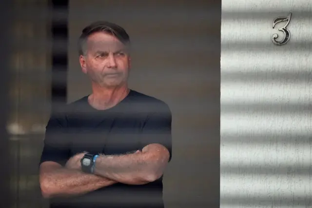 Bolsonaro de braços cruzados na frente de casa; é possível ver rastro de grades
