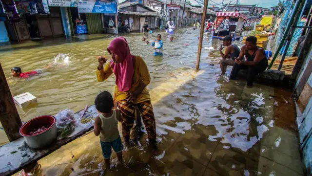 Alguns adultos se sentam à beira da estrada com os pés submersos na água, enquanto crianças caminham e nadam pela rua alagada no norte de Jacarta, em novembro de 2024
