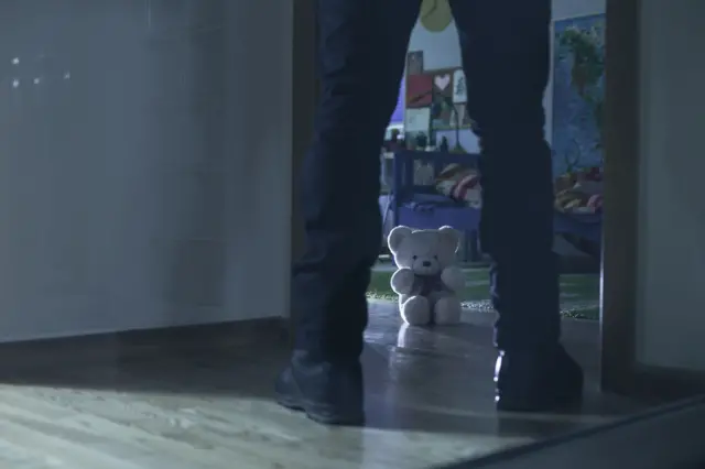 Напівтемний домашній коридор, у якому видно ноги дорослої людини, що стоїть у дверному прорізі. Попереду, у дитячій кімнаті, на підлозі сидить плюшевий білий ведмедик, освітлений слабким світлом.