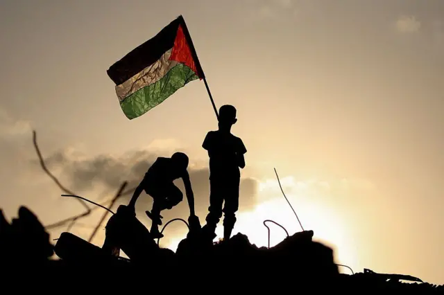 د غزې په مرکزي برخه کې د بوریج کډوالو په کمپ کې دوه ماشومان د فلسطین بیرغ په داسې حال کې پورته کوي چې د یوې ویجاړې شوې ودانۍ په کنډوالو ولاړ دي، د سپتمبر ۲۲، ۲۰۲۵.