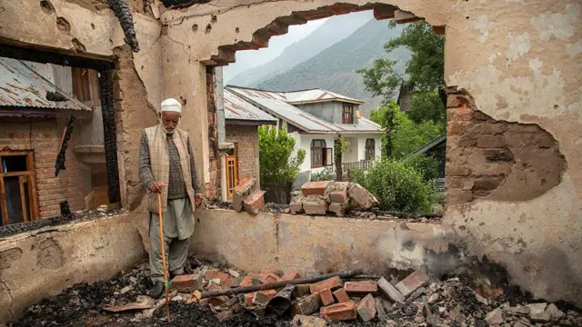 An elderly man stands inside the burned-out shell of his home, leaning on a wooden walking stick and staring quietly at the debris around him. He wears a white skullcap, a grey sweater beneath a beige vest and loose traditional trousers. The room is gutted and blackened, its walls cracked.