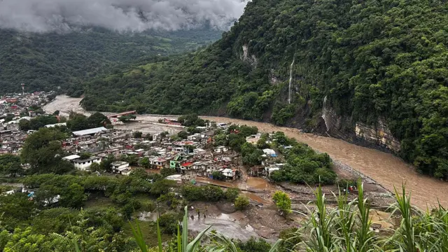 Vista general de una aldea al pie de una montaña, con casas de diferentes colores, está inundada por un río desbordado. En el fondo se ven nubes grises