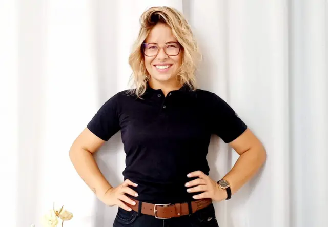 Rafaela Barbosa, mãe de um estudante que estuda em colégio cívico-militar, em um retrato; ela usa camisa preta, cinto e está com os cabelos soltos; o fundo é branco