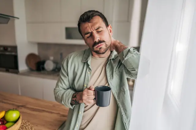 Pencere önünde elinde kahve fincanı ile bir adam. Diğer eliyle boynunu tutuyor. Gözleri kapalı.