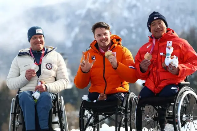 車いすに座った3人が横並びになり、手で持ったメダルを胸の前で見せている。上半身のウエアは、鈴木は赤、カンプスフルールはオレンジ、ペデシェンは白。全員笑顔。カンプスフルールは右手の指を3本立てている。背景には雪が残る山が見える