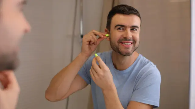 mężczyzna po czterdziestce używa zatyczek do uszu, aby uniknąć stresu związanego z hałaśliwymi sytuacjami. Patrzy na swoje odbicie w lustrze z uśmiechem.