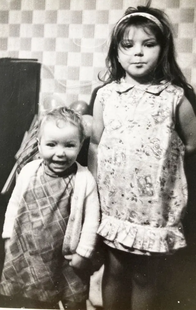 Uma fotografia em preto e branco de duas meninas dentro de casa. A mais velha está em pé usando um vestido estampado com uma tiara, enquanto a mais nova está ao lado, sorrindo, vestindo um conjunto xadrez e um cardigã. Atrás delas há uma parede com um papel de parede de padrão quadrado distinto e alguns balões