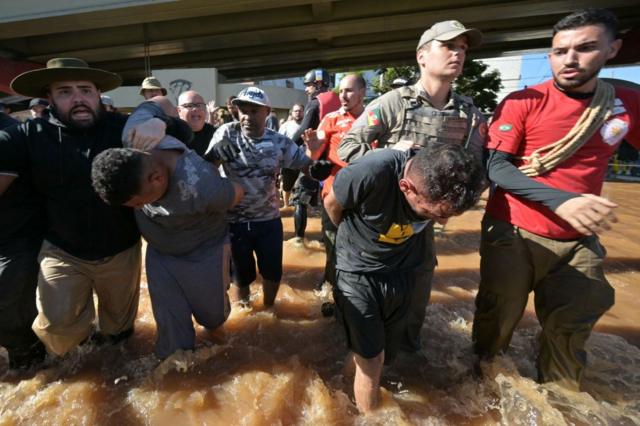 Pessoas e policiais em volta de dois homens carregados em posição curvada, com água nos joelhos