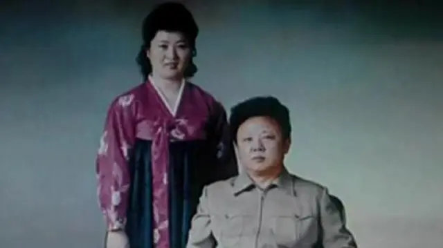 Ko Yong Hui, in a purple and green Korean dress stands on the left. Kim Jong Il sits besides her 