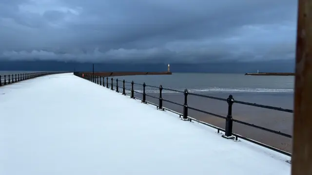 Vista del muelle curvo de Roker, cubierto por una capa de nieve intacta. El faro se alza al final del muelle, a la derecha. El cielo se ve oscuro y nublado, y el mar, al fondo, está en calma.