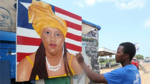 Wani mutum a gefen titi yana zana hoton wata mata a jikin tutar Liberia.