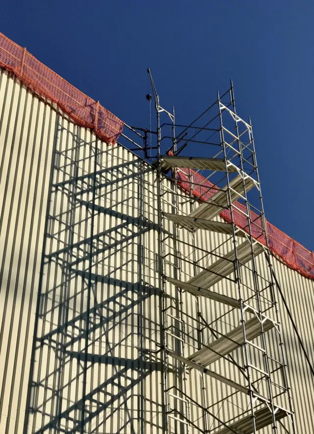 Scaffolding rises up along the side of a building and is framed against a blue sky