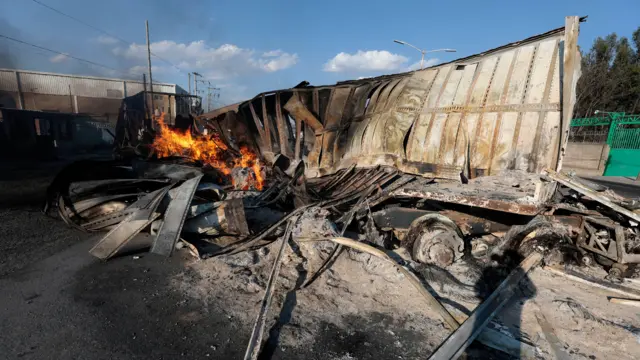 The hulk of a lorry on fire among rubble. A building and telegraph cables are nearby on a sunny day with blue skies.