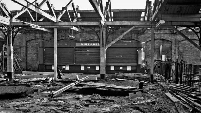 Una imagen en blanco y negro del vestíbulo interior de la estación de tren de Lowestoft muestra la demolición del tejado. Hay escombros y vigas apiladas en el suelo. Al fondo, una pared de ladrillos con una fachada tapiada con la inscripción «Quiosco Mullanes» sobre las ventanas tapiadas.