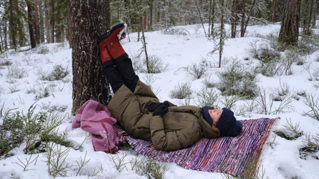 زنی روی برف‌ها در جنگلی دراز کشیده و پاهایش را بالا برده و به درختی تکیه داده است