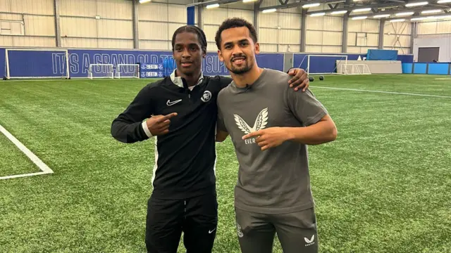 Kevar March-Mckenzie porte un survêtement Nike noir et désigne Iliman Ndiaye, qui se tient à côté de lui sur un terrain de football en salle. Ndiaye, vêtu d'un t-shirt gris et d'un bas de survêtement, désigne Kevar du doigt.
