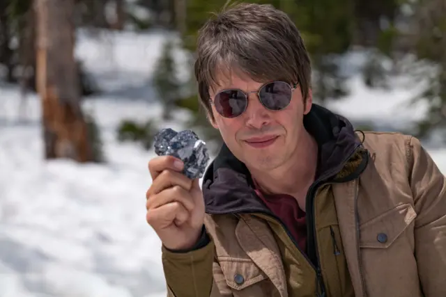 O Prof. Brian Cox posa para foto vestindo uma jaqueta marrom e óculos escuros. Ele segura uma pedra prateada feita de sulfeto de chumbo