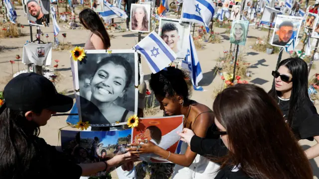 Pessoas agachadas prestam homenagem a vítimas com fotos e bandeiras de Israel em área aberta, com aparência de deserto