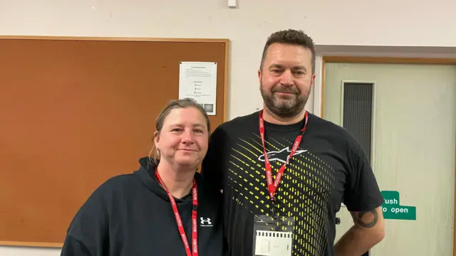 Una mujer con una sudadera negra con capucha está de pie junto a un hombre con una camiseta negra con un estampado amarillo. Ella lleva el pelo rubio oscuro recogido hacia atrás, mientras que él tiene el pelo corto y oscuro, y barba oscura.