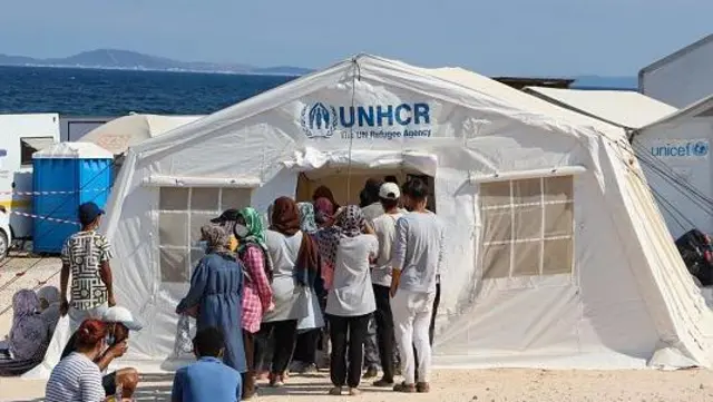 گروهی از پناهجویان در مقابل خیمه کمیسیاریای عالی پناهندگان ملل متحد در پس زمینه تصویر دریا دیده میشود