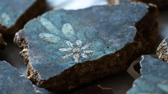 Pequeña pieza de pared de yeso con fondo negro, hojas de un azul más claro y una margarita blanca formada por un círculo central blanco y siete pétalos. También se pueden ver algunos otros fragmentos de yeso en los bordes de la imagen.