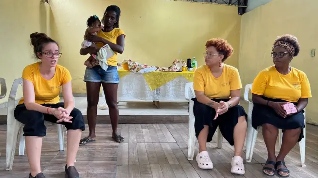Uma mulher em pé com bebê no colo e outras três mulheres sentadas em roda