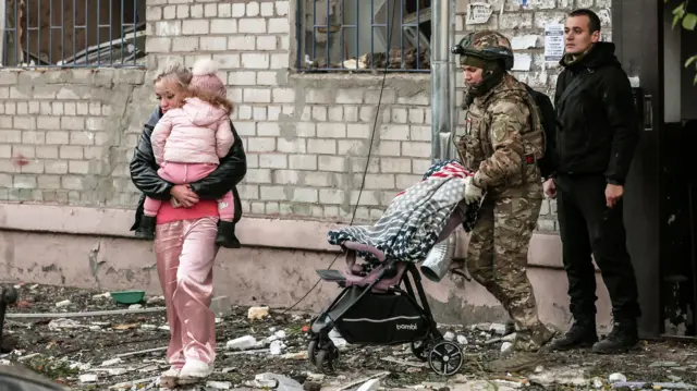 Una mujer, con un niño vestido con pantalones, chaqueta y gorro de lana rosas, sale de un edificio con escombros en el suelo. Un soldado uniformado empuja un cochecito y un hombre permanece de pie junto a la puerta del edificio.