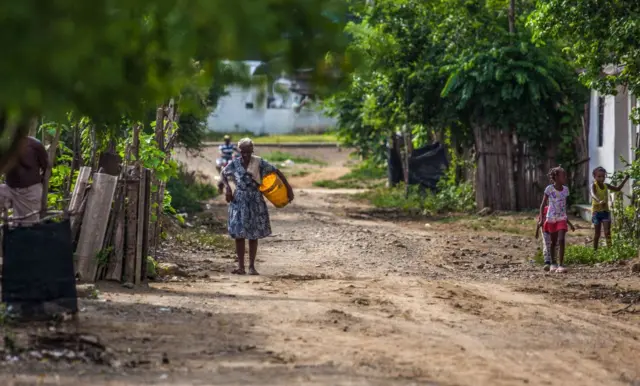 Una mujer afrodescendiente camina por una calle de tierra cargando un balde amarillo. Hay otros niños en la calle y un hombre en bicicleta en el fondo. Hay frondosa vegetación a lado y lado.