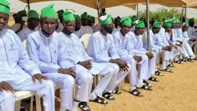 A group of repentant terrorists wearing white garments and yellow camps as dem dey pass out of di rehabilitation programme wey govment organise for dem.