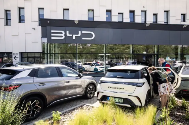 Egy budapesti BYD márkakereskedés előtt több elektromos autó áll. Két ember egy fehér autó utasterét nézi a nyitott ajtónál, a háttérben a szalon üvegfelülete.
