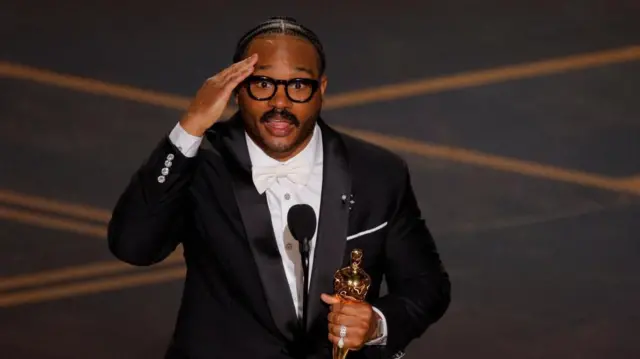 黒い上着と白い蝶ネクタイ姿で壇上で、オスカー像を持ったクーグラー監督が、額に片手をあてながら話している