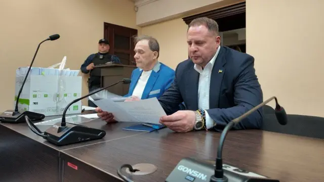 Ермак (справа) с адвокатом за столом в зале суда, у него в руках бумаги, рядом с адвокатом стоит наполненная бумагами коробка 