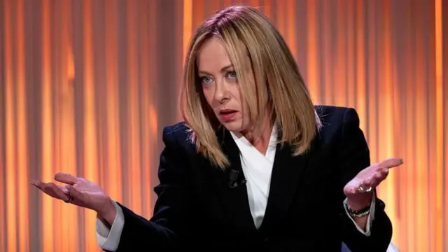 Siyah ceket ve beyaz bluz giymiş Meloni turuncu bir perde önünde iki elini iki yana açmış bir halde bakıyor