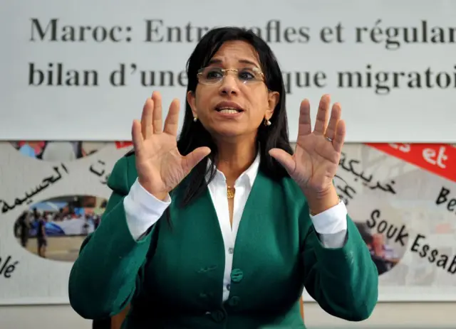 Amina Bouayach en veste verte avec chemise blanche, elle lève les deux mains et parle, en arrière-plan une affiche avec des écritures.
