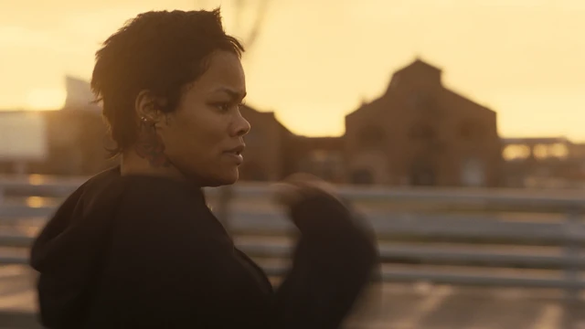 Жінка стоїть на вулиці під теплим, золотистим вечірнім світлом. Удалині видно розмиті контури великих будівель із характерними арками та вікнами.