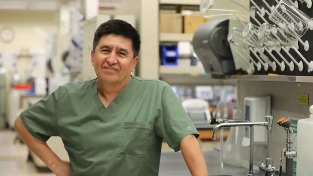 Ọkunrin yii wọ aṣọ alawọ ewe, o duro sinu yara ayẹwo kan ti awọn irinṣẹ wa nibẹ. O n rẹrin-in bo ṣe n wo oju Kamẹra.