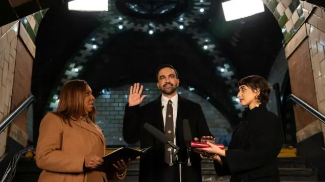 レンガ造りの建物の中で、黒いコートを着たマムダニ新市長が右手を上げて宣誓している。