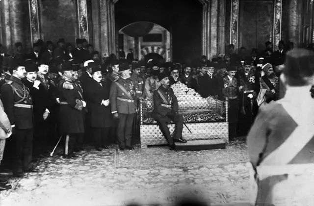 En un gran salón, está Mehmed VI sentado solo en un sofá y detrás de él hay varios hombres que parecen funcionarios
