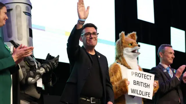 「ビンフェイス（顔がごみ缶）伯爵」ことコメディアンのジョナサン・ハーヴィー氏や、「イギリスの野生動物を保護しろ」党の候補などと並んだバーナム氏が、右手を挙げて笑顔で挨拶している