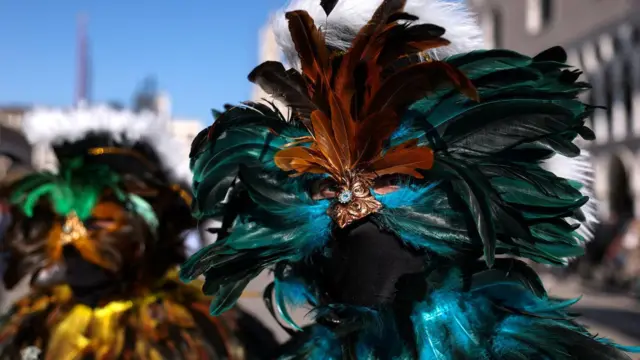 Muškarac drži masku napravljenu od plavo-zelenih pera, vide mu se samo oči