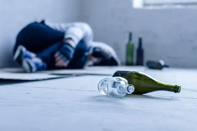 На підлозі порожнього приміщення лежать кілька порожніх скляних пляшок на передньому плані. У розмитому тлі біля стіни лежить людина, згорнувшись клубком.