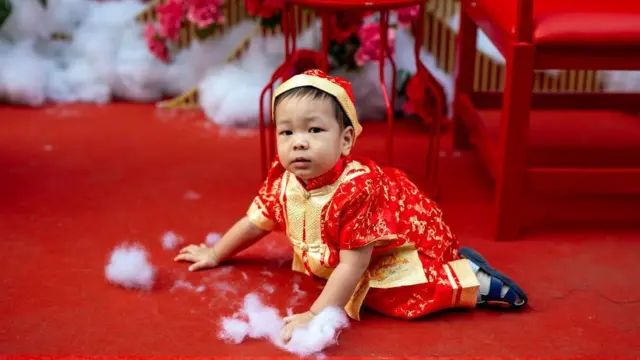 یک کودک چینی با لباس سنتی روی فرش سرخ رنگ که پنبه سفید زیر دستانش قرار داده شده است.