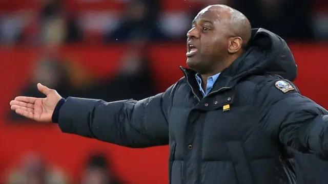 Patrick Vieira, que l'on voit du ventre jusqu'à la tête, vêtu d'une épaisse doudoune noire par-dessus une chemise bleu clair, tend le bras droit le long du corps et proteste lors d'un match de football. On aperçoit une tribune, floue, derrière lui, au loin.