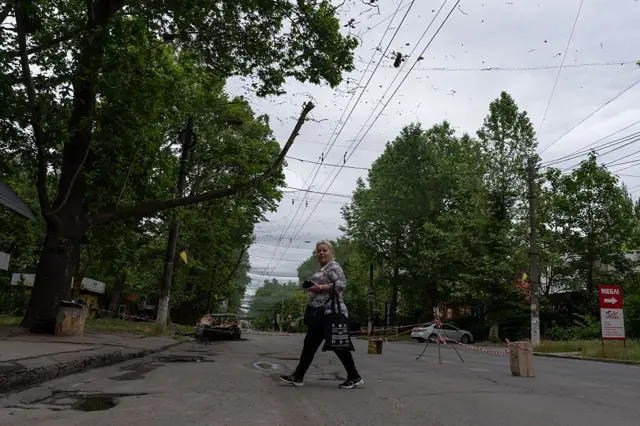 На міській вулиці, оточеній високими деревами, дорогу перегороджено червоною стрічкою. Над проїжджою частиною натягнута антидронова сітка: на ній висить невеликий безпілотник, далі на дорозі видно обгорілий автомобіль. Попереду просто попід дроном йде жінка з сумкою, дивлячись у камеру.